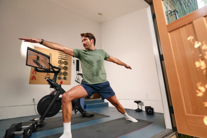 Ein Mann in verschwitzen Shirt steht auf einer Yogamatte von Peloton und befindet sich in der Krieger-Pose. Er folgt den Anweisungen eines Trainers auf dem Peloton Bike+, welches hinter ihm steht. Den Bildschirm hat er so gedreht, dass er alles sehen kann.