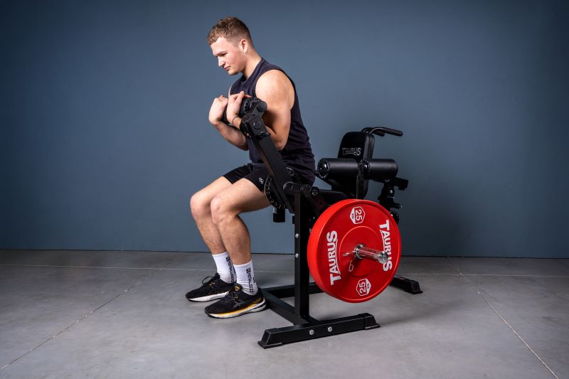 Ein junger Mann sitzt auf dem 4-in-1 Multitrainer von Taurus und hält vor der Brust die Rolle und führt mit dieser Crunches aus. Das Gerät ist mit einer Hantelscheibe von Taurus belegt.